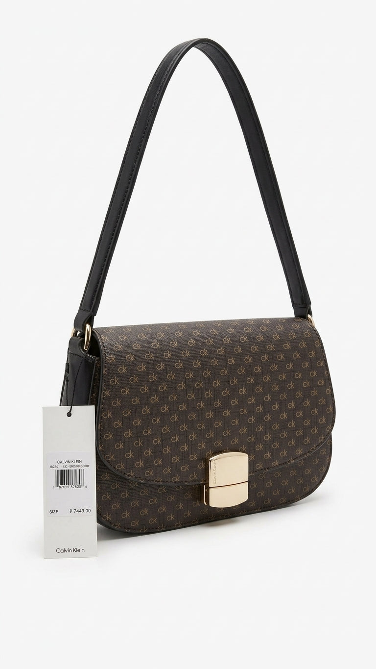 A dark brown monogram shoulder bag with a black leather strap and a gold-tone metal clasp, shown with its white Calvin Klein product tag on a clean white background.